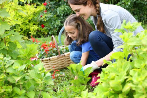 A woman and a young girl are engaged in gardening within a lush, well-maintained backyard. They are crouched down amidst vibrant green foliage, including broad-leaved plants and flowering bushes, with some red flowers visible in the background. The woman has long brown hair styled in a braid and is wearing a light grey top and blue jeans, while the girl is dressed in a blue top and jeans. They are focused on the task of planting or tending to plants, with the girl holding a wicker basket, possibly for collecting garden produce or weeds. The garden area features a neatly trimmed lawn with dense, healthy grass, bordered by a variety of shrubs and flowering plants, creating a cheerful and natural outdoor environment suitable for family gardening activities. The overall scene is well-lit with natural daylight, highlighting the lush greenery and colorful blooms, demonstrating typical garden maintenance and outdoor care activities that a professional gardening service like Hedge Trimming Pimlico may support for local residents in Pimlico.
