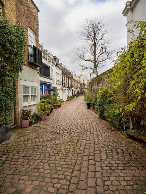 A narrow, winding cobblestone alleyway in Pimlico mews lined with picturesque terraced houses featuring white and pastel-colored facades, with small front gardens decorated with potted plants and shru