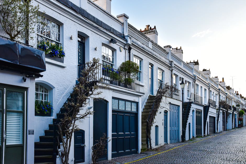 A narrow, winding cobblestone alleyway in Pimlico mews lined with picturesque terraced houses featuring white and pastel-colored facades, with small front gardens decorated with potted plants and shru
